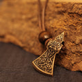 The Viking Axe Brass Pendant showcases intricate knotwork and a rune symbol, highlighted by a wooden bead on the cord. Resting on a coarse brown surface, this Norse jewelry makes a striking addition to any collection.