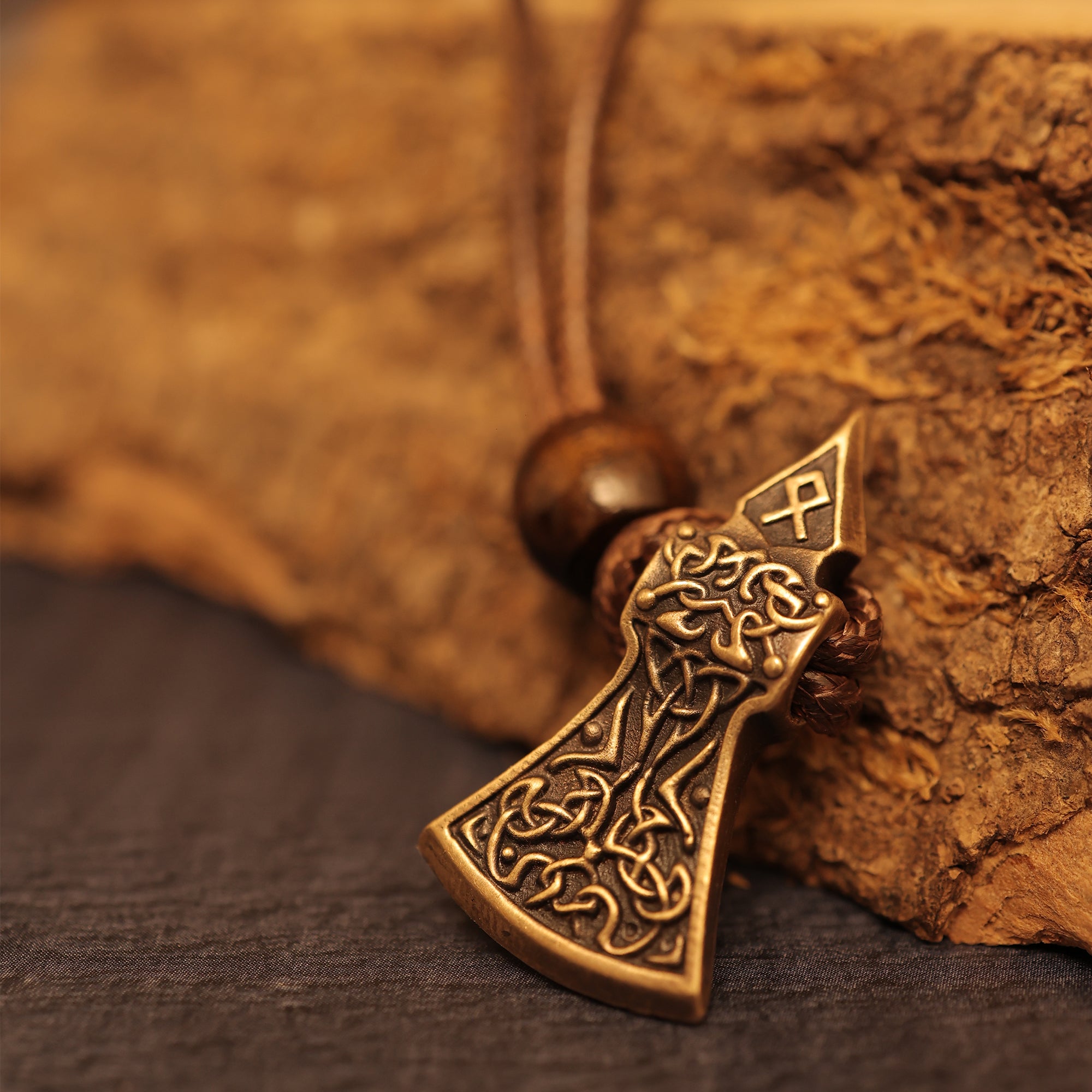The Viking Axe Brass Pendant showcases intricate knotwork and a rune symbol, highlighted by a wooden bead on the cord. Resting on a coarse brown surface, this Norse jewelry makes a striking addition to any collection.