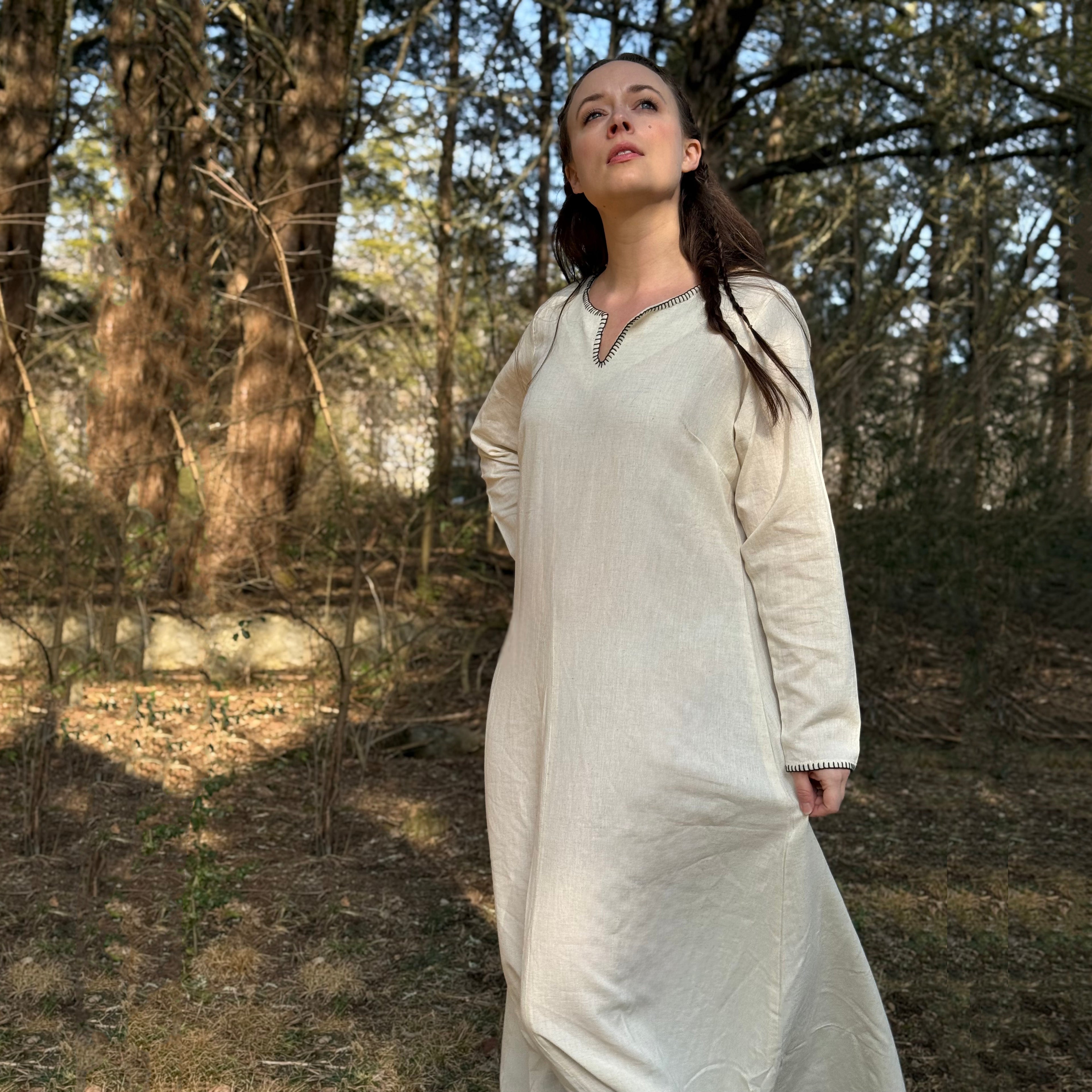 A woman stands outdoors in a sunlit forest, wearing the Viking Thorndale Dress- Ladies, a long, light-colored gown with black trim. She gazes upward serenely, one hand at her side and the other behind her back.