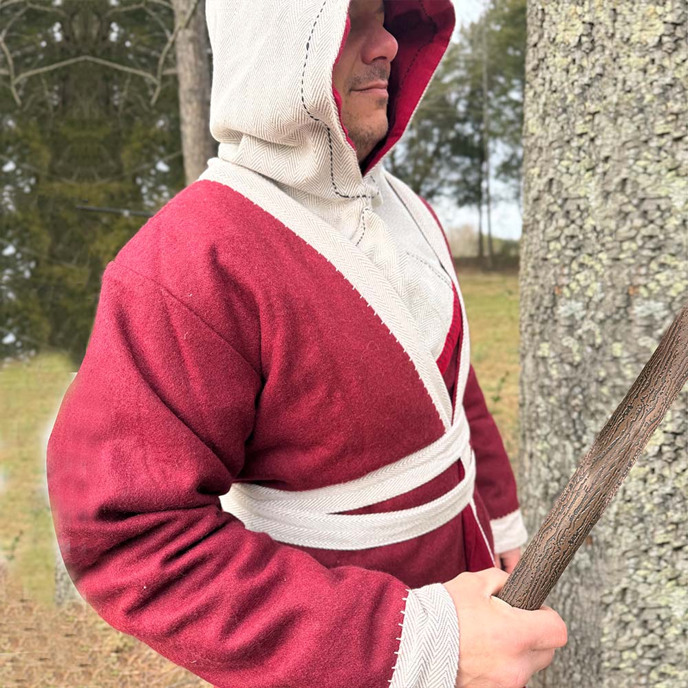 A person wearing the Viking Wool Coat - Men's in red and cream stands outdoors beside a tree and holds a wooden staff, with trees and grass in the background creating a forested atmosphere.