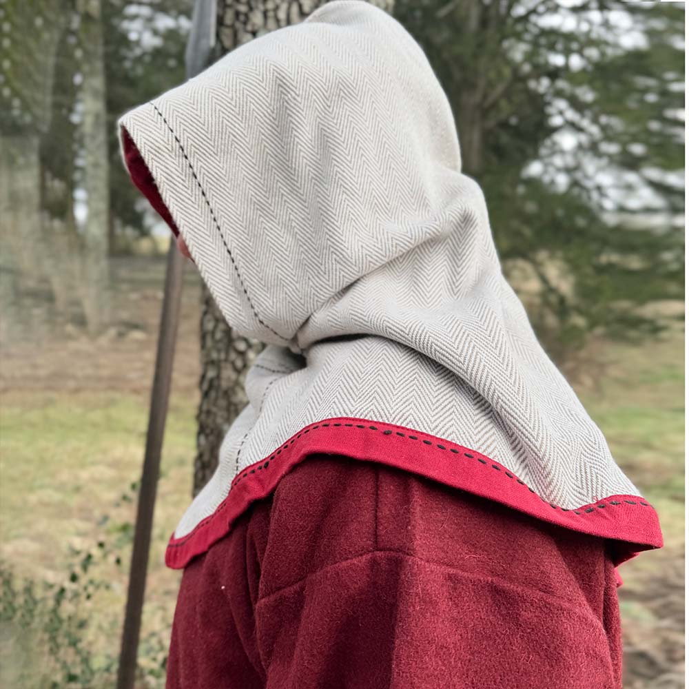 A person wears a beige Reversible Viking Hood, Skjoldehamn with red trim and a red cloak, standing outdoors by a tree against a blurred natural background—ideal for medieval reenactment.