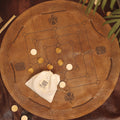 A round leather Nine Men's Morris board with a Celtic knot border and four symbols, featuring light and dark wooden pieces. A bag labeled MILL GAME rests on the board; green leaves sit at the top right.