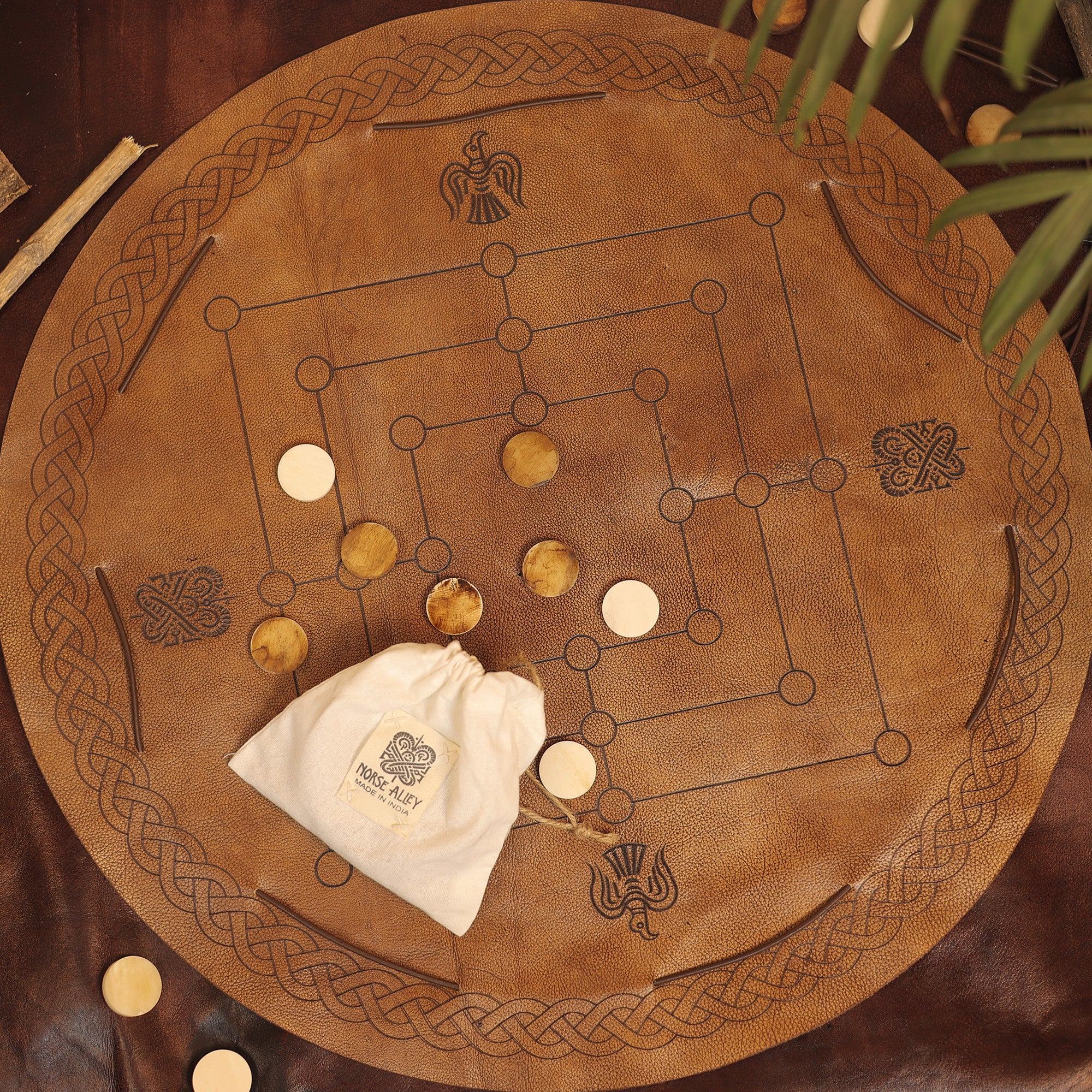 A round leather Nine Men's Morris board with a Celtic knot border and four symbols, featuring light and dark wooden pieces. A bag labeled MILL GAME rests on the board; green leaves sit at the top right.
