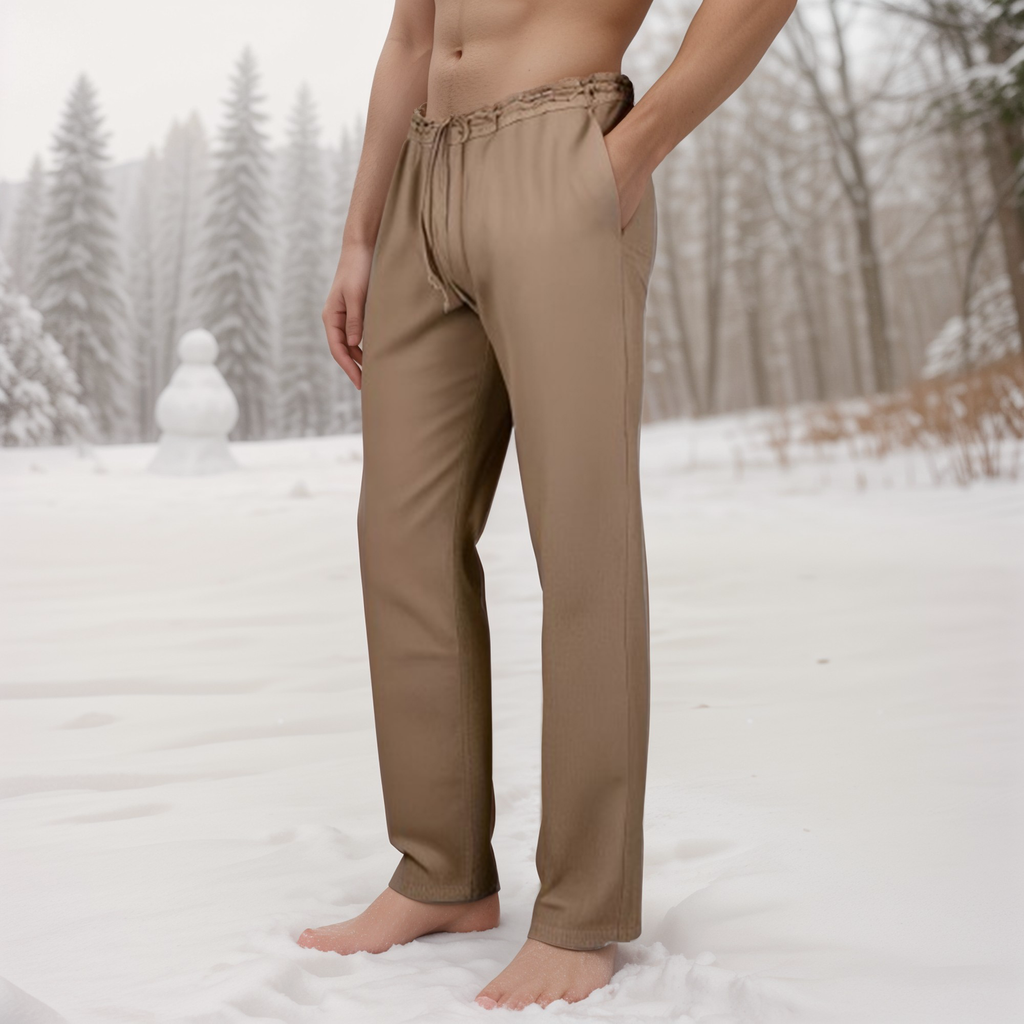 A barefoot man wearing Warrior Pants - Men's stands in the snow, with a snowy forest and a snowman in the background.