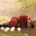 Two Handcrafted Dark Red Leather Dice Cups—one upright, one on its side with heart stitching—rest beside a rock, five white dice, and green leaves, making them standout tabletop RPG accessories against a neutral background.