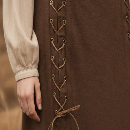 A close-up of a hand resting by their side, featuring the Medieval Skirt- Ladies in brown with vertical lace-up details and metal eyelets, paired with a beige long-sleeved blouse.