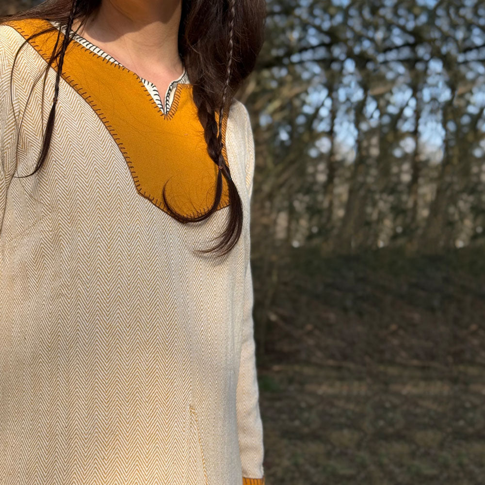 A woman stands outdoors by trees, sunlight casting soft shadows, wearing the Nordic Runeweave Viking Dress-Ladies—a cream outfit with mustard yellow neckline and cuffs that reflects the elegance of Norse craftsmanship.