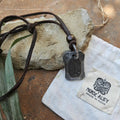 The Viking Rune Pendant Isa, featuring a metallic Isa rune on a brown leather cord, is displayed on a stone beside a Norse Alley pouch and a green leaf, highlighting this striking Norse necklace.