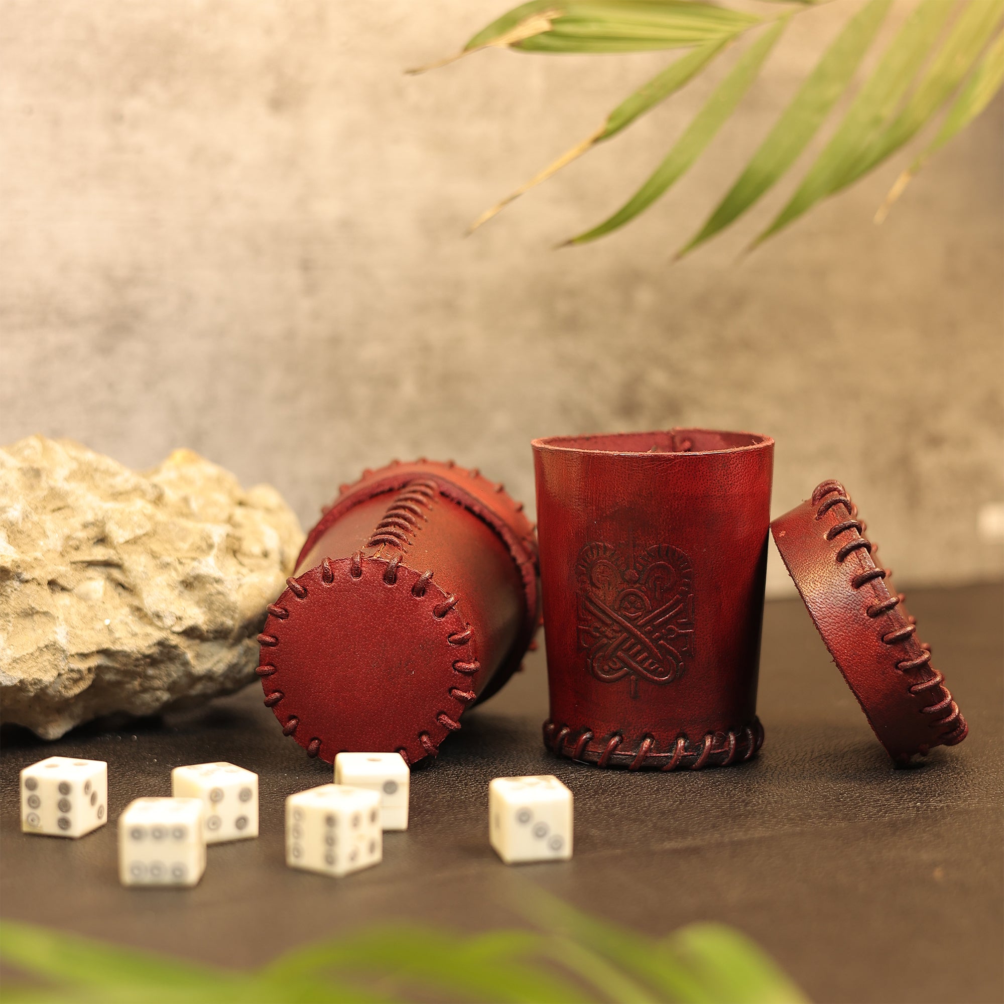 Two Handcrafted Dark Red Leather Dice Cups—one upright, one on its side—sit beside a rock. Six white dice with black dots are scattered in front, green leaves above. Perfect tabletop RPG accessories.