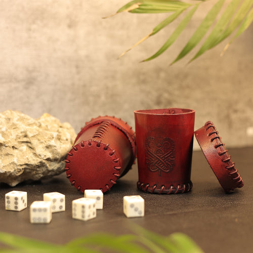 Two Handcrafted Dark Red Leather Dice Cups—one upright, one on its side—sit beside a rock. Six white dice with black dots are scattered in front, green leaves above. Perfect tabletop RPG accessories.