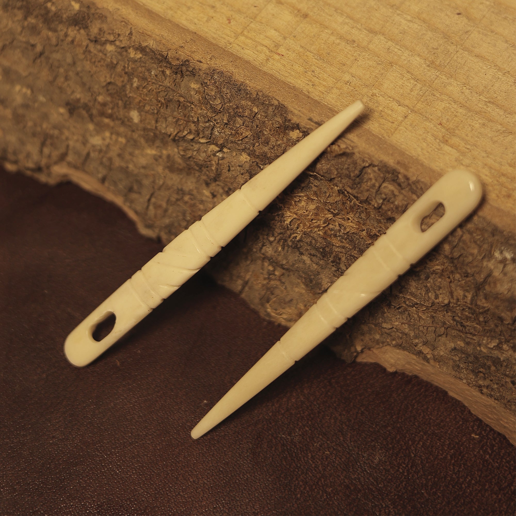 A pair of handcrafted Viking style bone knitting needles with pointed ends and oval holes rests on a rustic wooden surface and brown leather.
