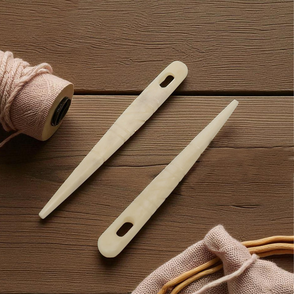 A pair of handcrafted Viking style Bone Knitting Needles lies parallel on a wooden surface, surrounded by pink yarn and a spool of thread.