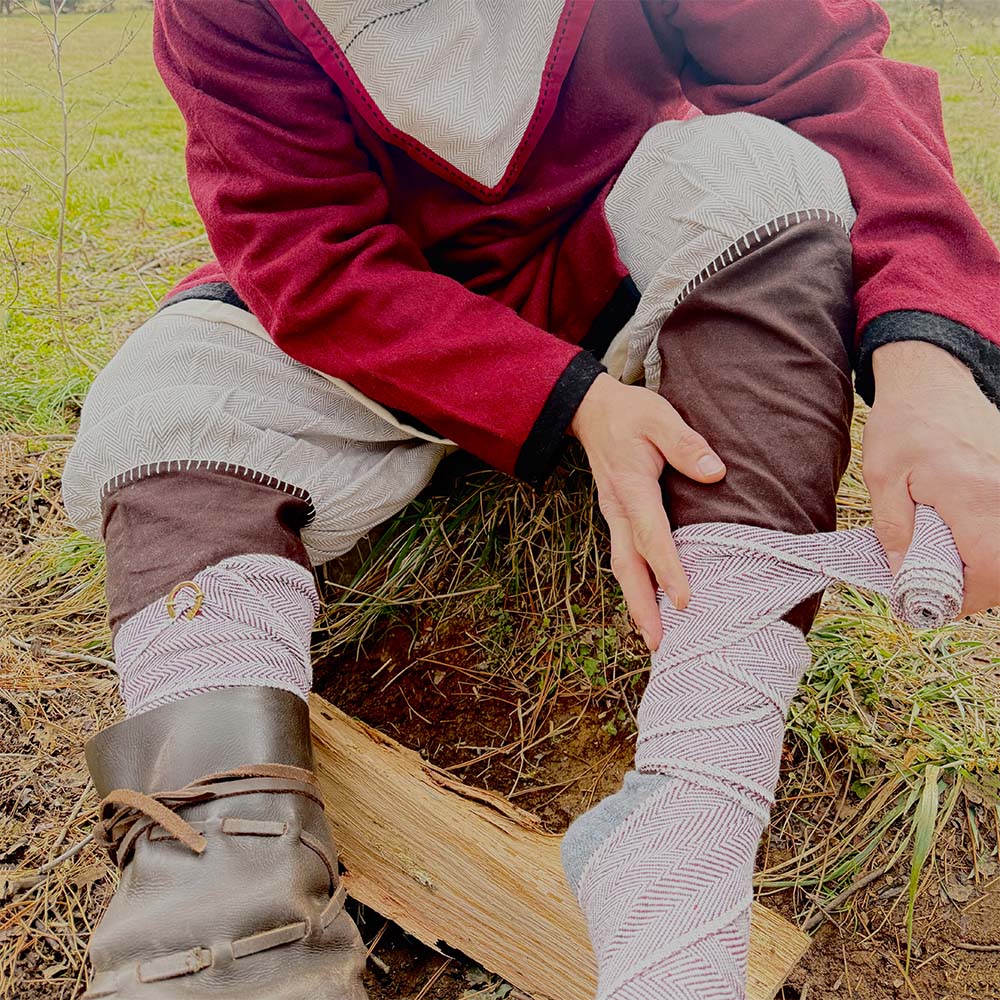 A person in historical clothing sits on grass, wrapping Herringbone Viking Leg Wraps (Winingas) over brown pants and boots, preparing for a reenactment or outdoor activity.
