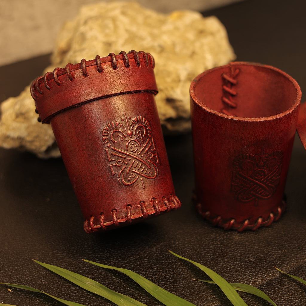 Two Handcrafted Dark Red Leather Dice Cups with stitched edges and embossed Viking knot designs rest on a black surface, surrounded by rocks and green leaves—ideal accessories for tabletop RPG sessions.
