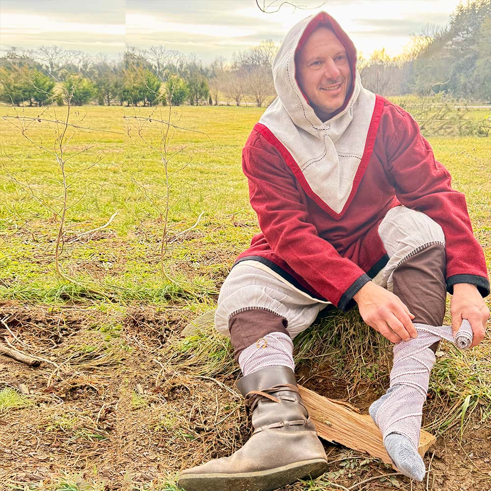 Wearing a red tunic, gray hood, brown pants, and leather boots, a person sits on the grass beneath a cloudy sky, smiling as they roll up Herringbone Viking Leg Wraps (Winingas) amid trees for an authentic medieval look.