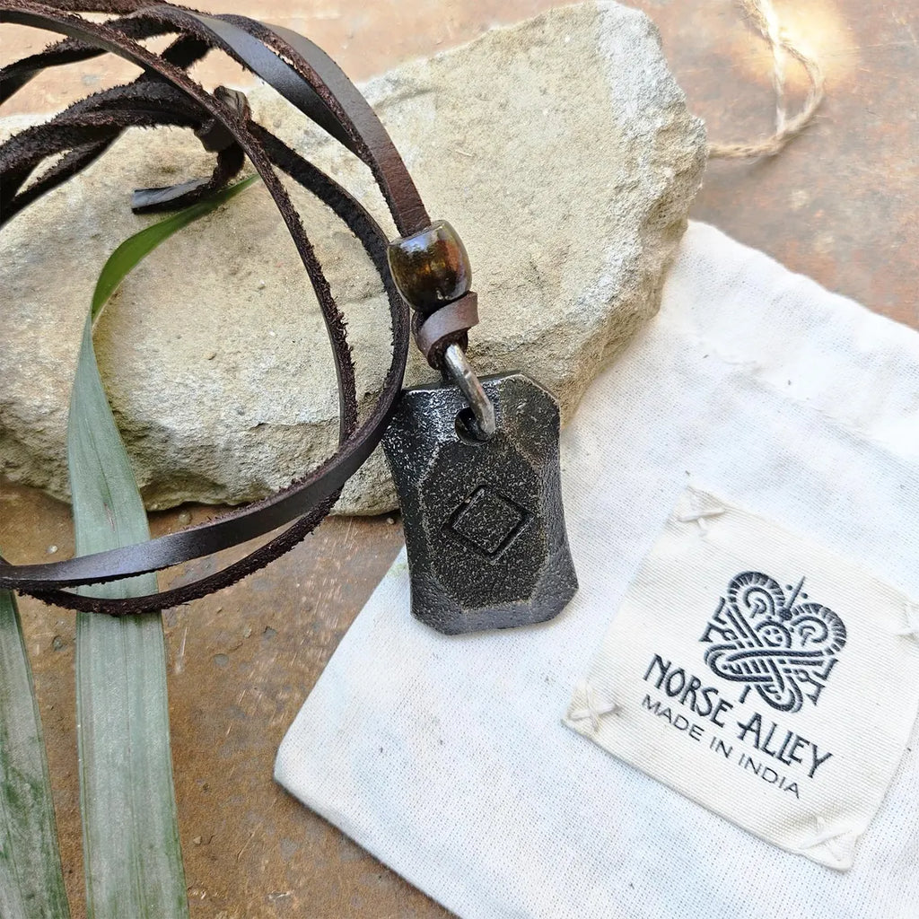 A black Viking Rune Pendant Ingwaz on a leather cord sits on a rock next to a folded white pouch labeled NORSE ALLEY MADE IN INDIA with Norse design, while a green leaf is nearby.
