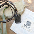 A black Viking Rune Pendant Ingwaz on a leather cord sits on a rock next to a folded white pouch labeled NORSE ALLEY MADE IN INDIA with Norse design, while a green leaf is nearby.