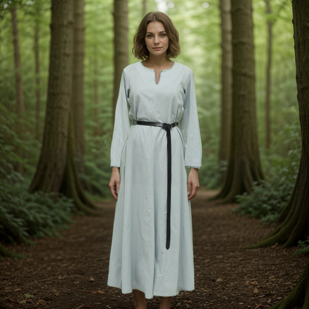 A woman wearing the Viking Thorndale Dress – Ladies in light blue with a black belt stands on a forest path, surrounded by tall trees and lush greenery.