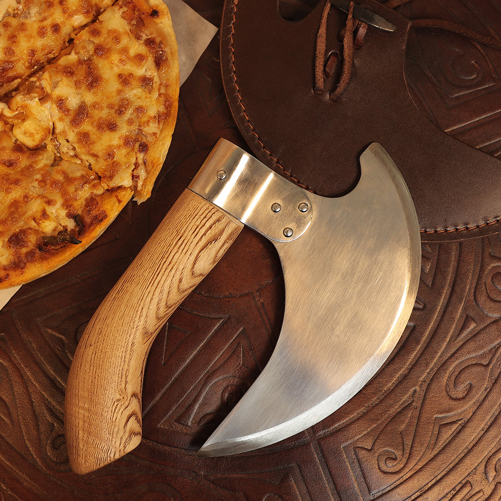 A close-up of a cheesy pizza beside the Pizza Axe with Brown Scabbard, featuring a stainless steel blade and wooden handle, resting on an intricately patterned surface with its leather scabbard nearby.