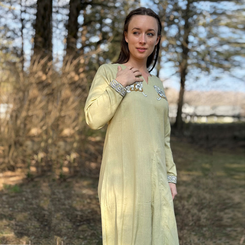 A woman in the Sleipnir Viking Dress—long, light-colored cotton-linen with decorative trim—stands outdoors among tall trees, sunlight filtering through as she looks to the side with one hand gently touching her chest.