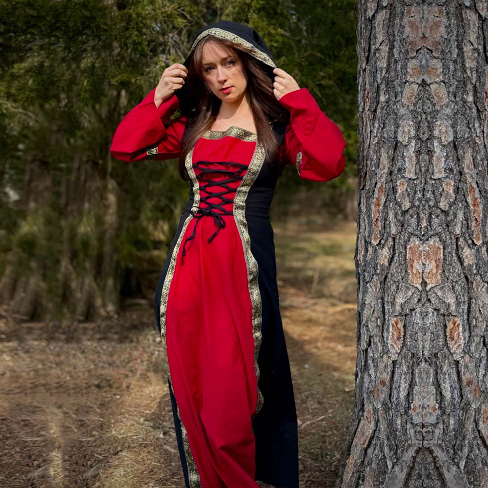A woman in the Dark Majesty Medieval Dress for ladies, featuring red and black fabric with gold trim, stands by a tree in a forest, lifting her hood—a perfect choice for LARP clothing or Renaissance fair costumes.
