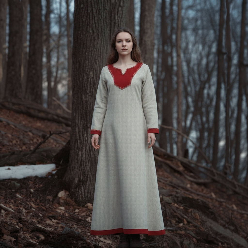 A woman wearing the Nordic Runeweave Viking Dress-Ladies, featuring red trim, stands in a serene, wintry forest with bare trees and patches of snow amid fallen leaves.