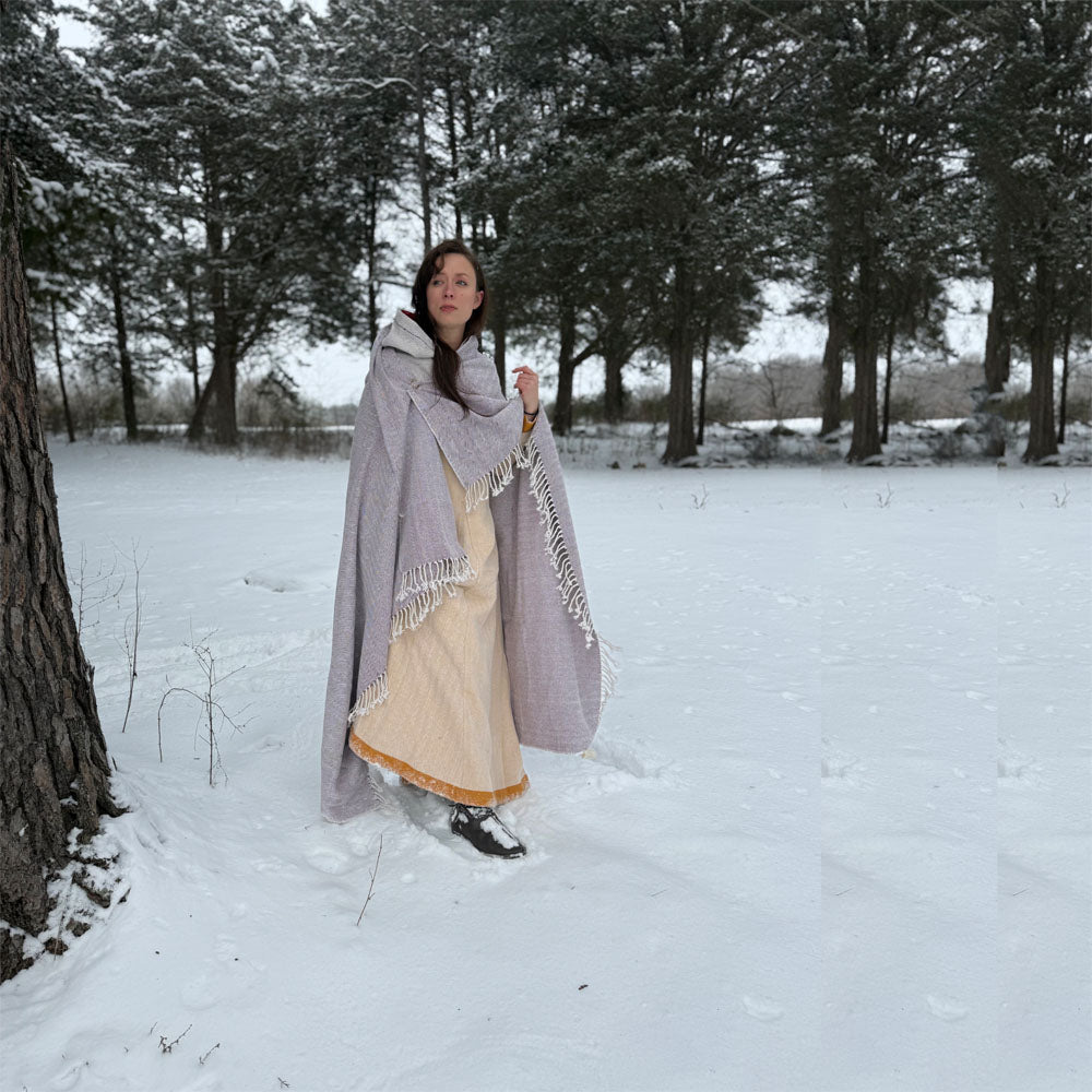 Wearing Odin's Viking Cape—Unisex, a woman in a pale dress with a fringed shawl stands in a snowy pine forest. She gazes aside as snow blankets the ground and trees, capturing the timeless spirit of Norse culture.