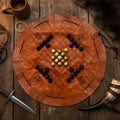 The Handcrafted Viking Hnefatafl Set features a circular leather board with dark and light hand-carved bone and horn pieces, displayed among medieval props like a horn, dagger, mug, fur, and leather mask on a wooden table.