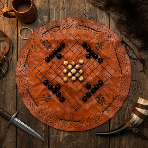 The Handcrafted Viking Hnefatafl Set features a circular leather board with dark and light hand-carved bone and horn pieces, displayed among medieval props like a horn, dagger, mug, fur, and leather mask on a wooden table.
