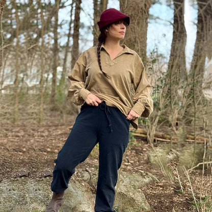 A woman wears the Warrior Shirt – Medieval Strength & Style- Unisex, paired with a red hat and black pants, standing on a rock in the woods, looking to the side with one hand on her hip and the other on her raised knee.