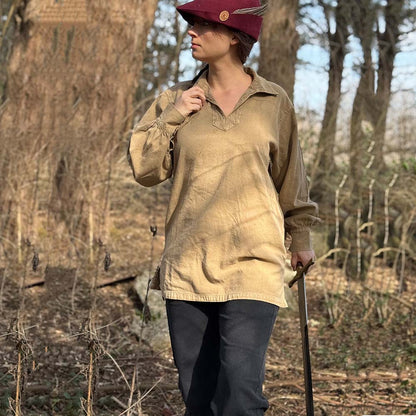 A person wears the Warrior Shirt – Medieval Strength & Style- Unisex, paired with black pants and a red hat, standing in a sunlit forest while holding a stick and gazing to the side.