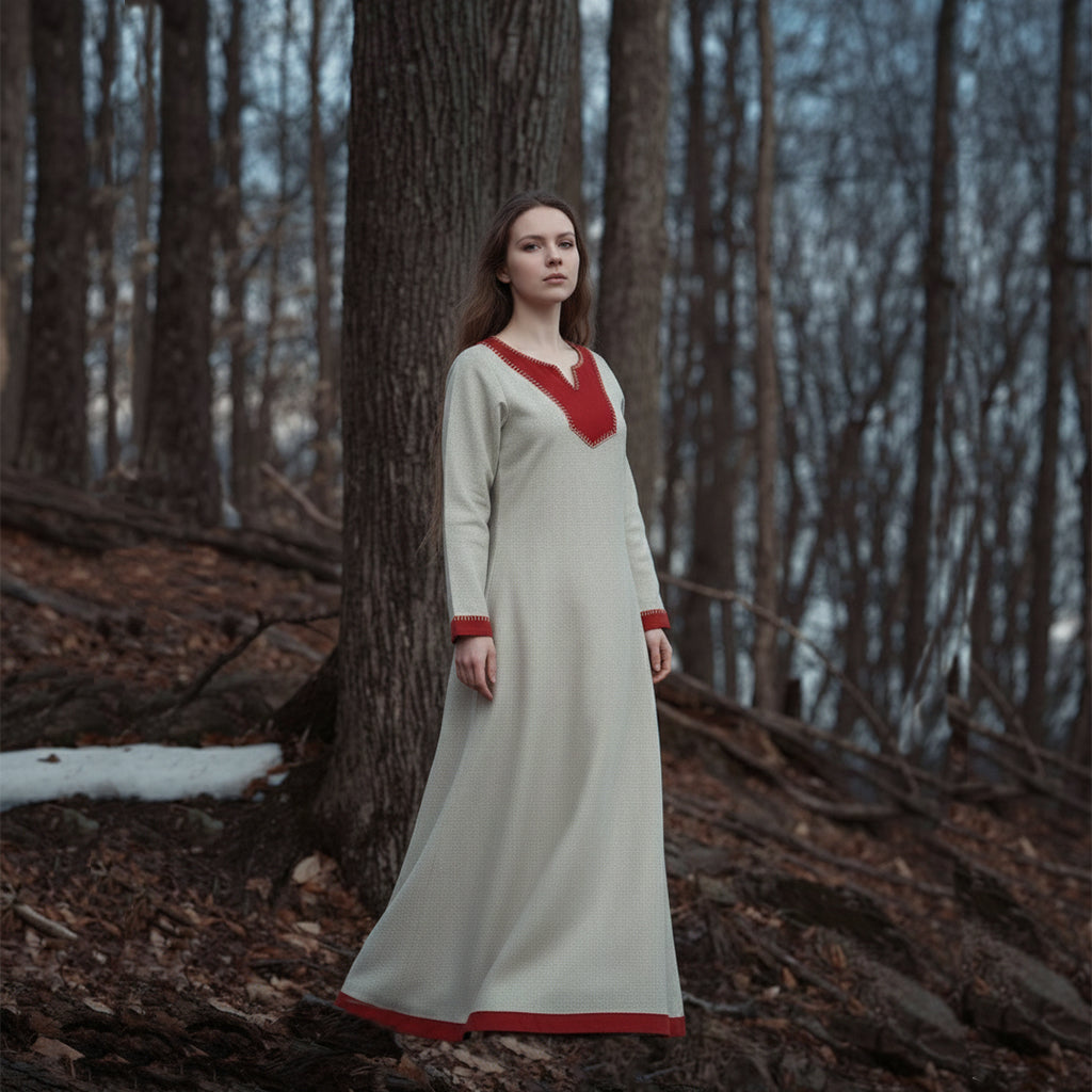 A woman wearing the Nordic Runeweave Viking Dress-Ladies, a long cream-colored gown with red accents and hand-embroidered Norse details, stands calmly in a snowy, leaf-covered forest of bare trees.