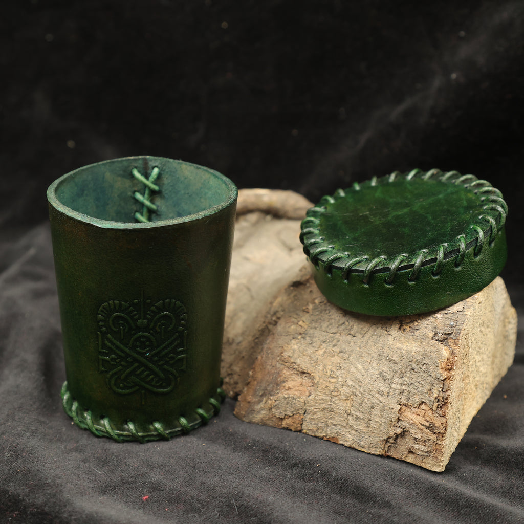 A handcrafted dark red leather dice cup with decorative stitching and a matching lid sits on wood, featuring an embossed design on its side—ideal for tabletop RPG accessories.