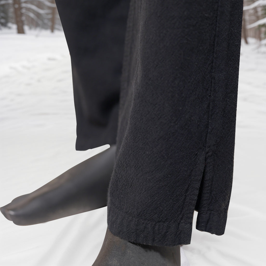 Close-up of a person standing barefoot on snow, wearing Warrior Pants- Men's in black. The setting features snow-covered ground and trees, highlighting the pants' Viking-inspired style suited for cold outdoor conditions.
