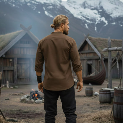 A blond-haired man in a bun stands before Viking wooden houses, a longship, and snowy mountains. Dressed in the Warrior Shirt – Medieval Strength & Style- Unisex and black pants, he evokes a historical Norse costume setting.