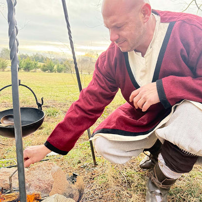 Wearing the Viking Woolen Tunic- Men's, a person crouches by an outdoor campfire with a black pot hanging over the flames on a metal tripod, set in a grassy field with trees behind.