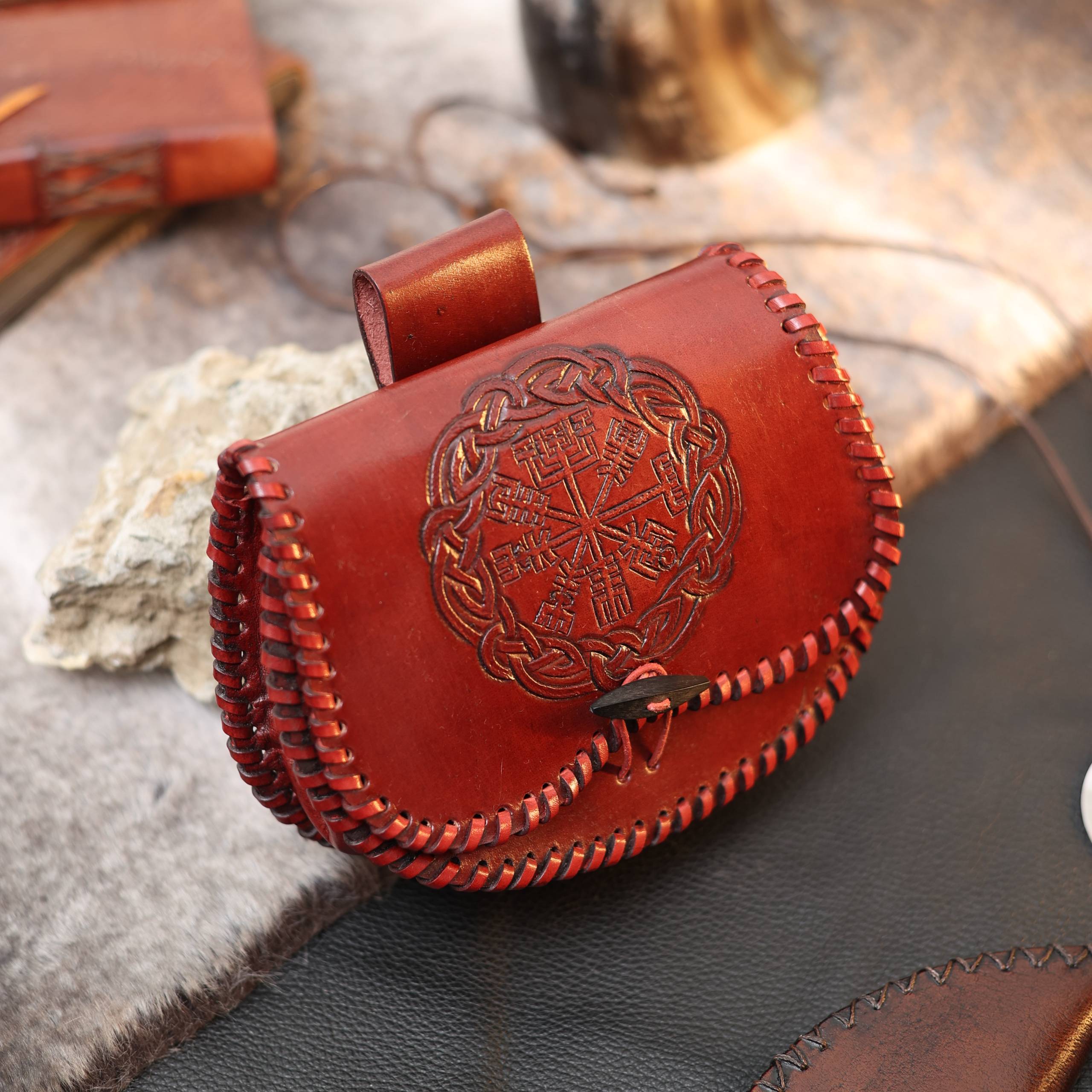 The Vegvisir leather Viking bag features reddish-brown leather, braided stitching, and an embossed Vegvisir symbol inspired by Norse mythology. Resting on fur and leather, it makes a unique gift for any Norse mythology enthusiast.