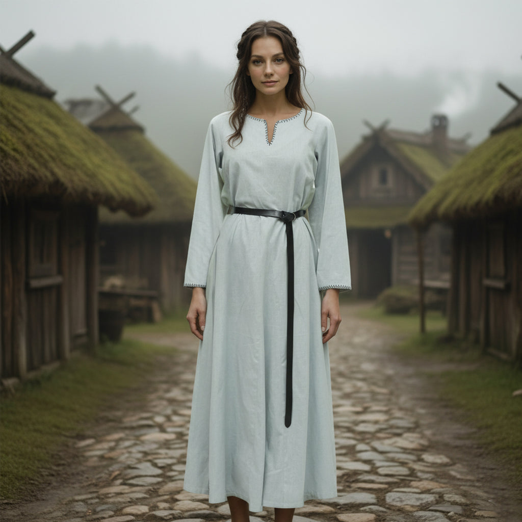 A woman in the Viking Thorndale Dress- Ladies, a simple long light blue gown with a black belt, stands on a misty cobblestone path in a rustic village of wooden houses and thatched roofs.
