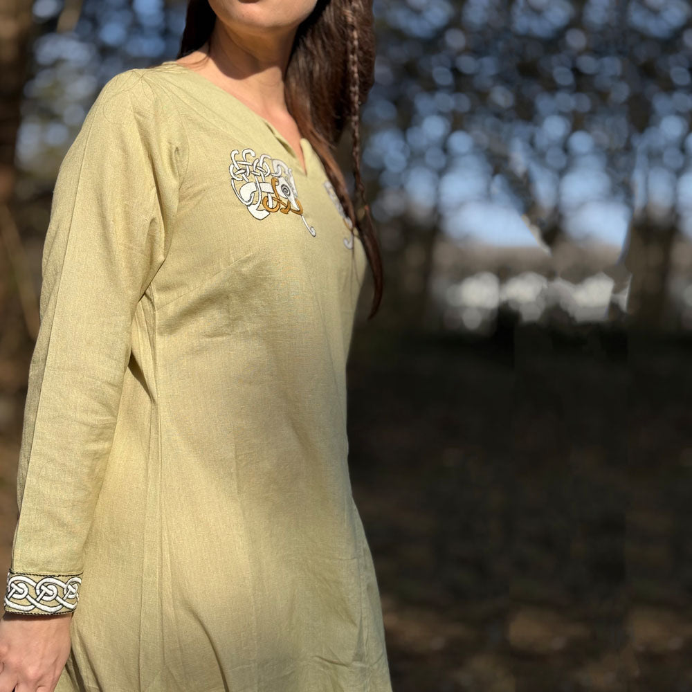 Someone stands outdoors in sunlight, surrounded by blurred trees, wearing the Sleipnir Viking Dress—Ladies: a light yellow cotton-linen dress with white and brown chest embroidery and patterned cuffs.