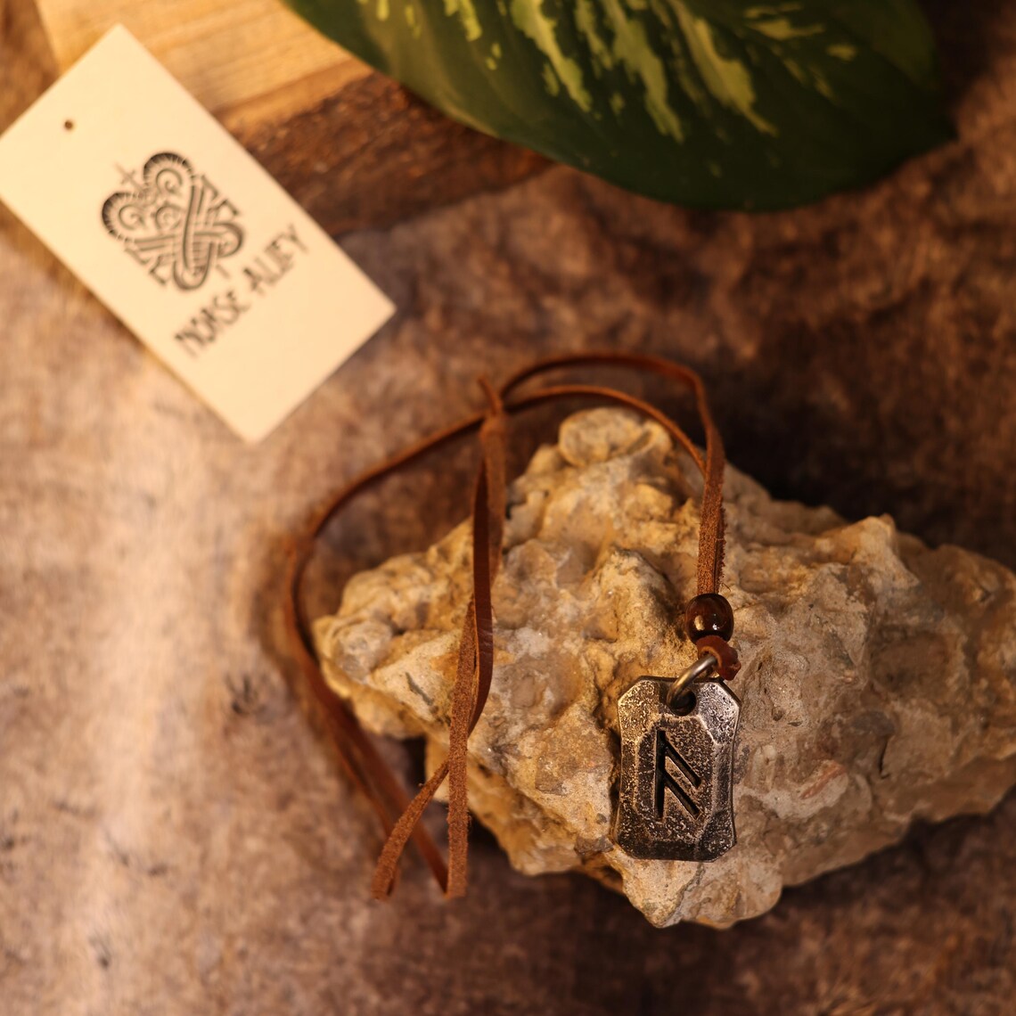 The Viking Rune Pendant Ansuz hangs from a brown leather cord on textured stone, accented by a leaf and a Norse Alfy tag. Warm lighting enhances the earthy, natural vibe of this rustic Viking jewelry scene.