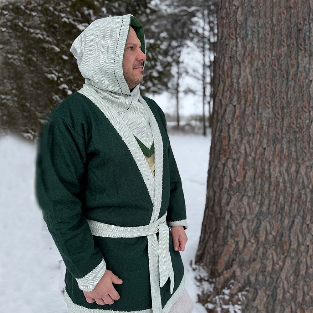A person in a green and light gray Viking Wool Coat - Men's stands by a large tree in a snowy forest, gazing into the distance.