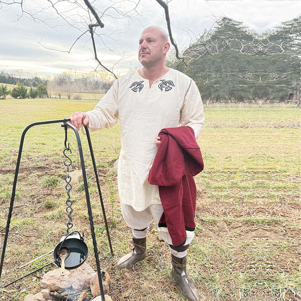 A person in historical clothing stands outdoors near a fire pit, holding the Odin's Raven Tunic- Men's, with grass, trees, and a cloudy sky in the background.