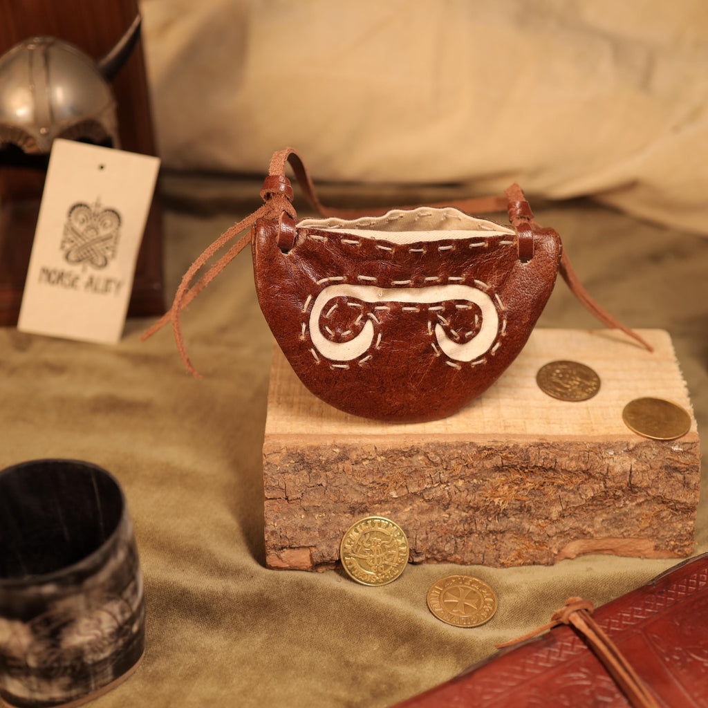 The Gokstad Wallet Viking Coin Pouch, a small brown handcrafted leather wallet with white stitching, rests on a wooden block amid gold coins and rustic décor, featuring a helmet and Viking-style tag in the background.