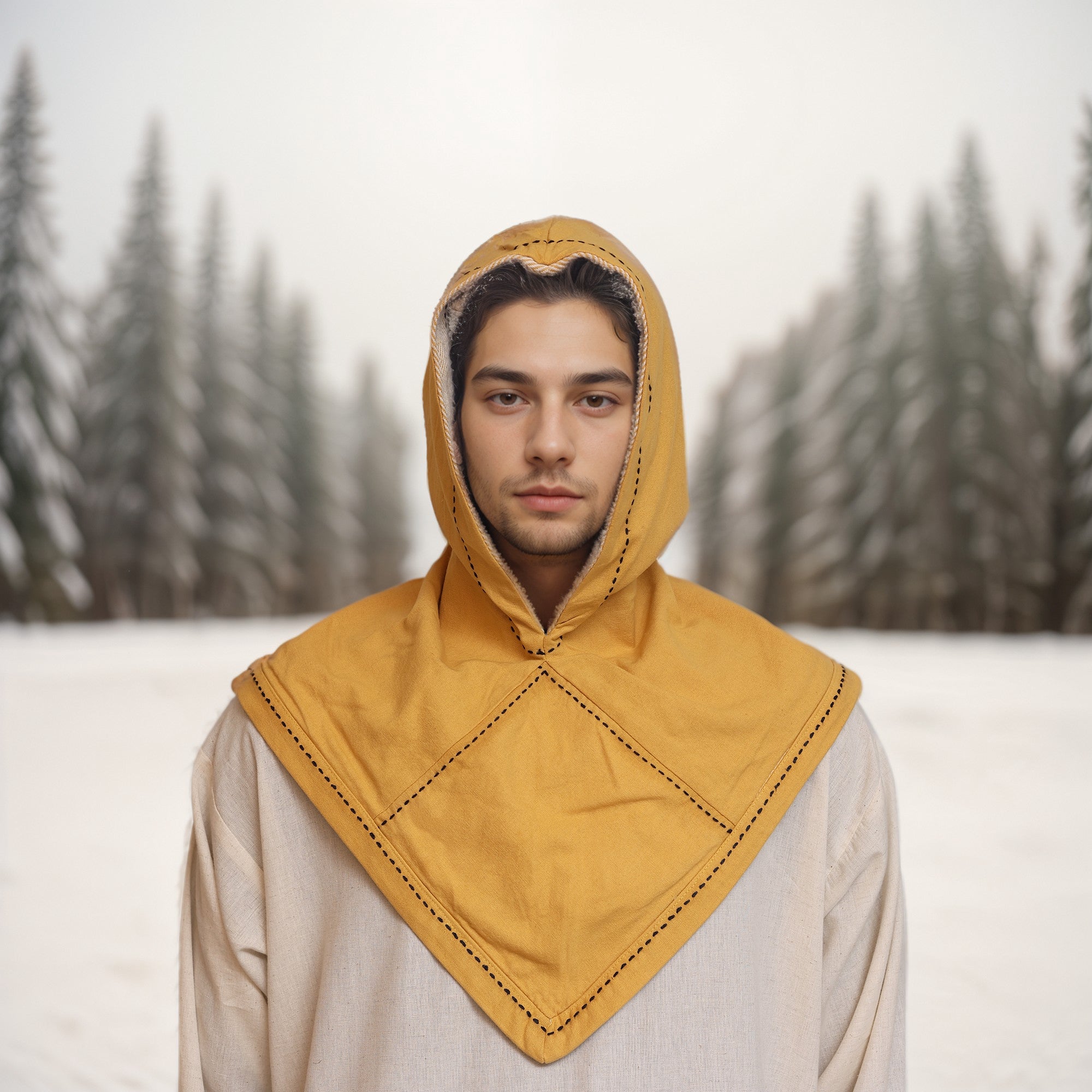 A young person wears a tan Reversible Viking Hood, Skjoldehamn, and a yellow medieval-style mantle while standing in a snowy forest with evergreen trees in the background.
