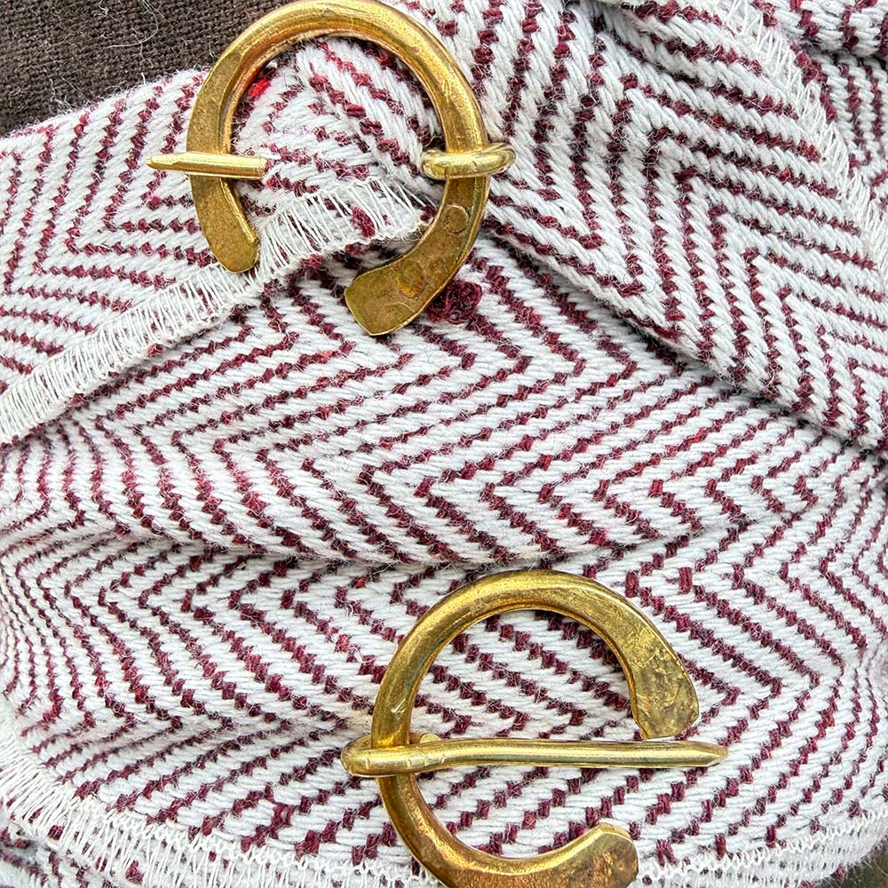 Two circular brass shawl pins rest on Herringbone Viking Leg Wraps (Winingas), which feature a red and white herringbone pattern and frayed edges, inspired by traditional medieval Viking legwear.