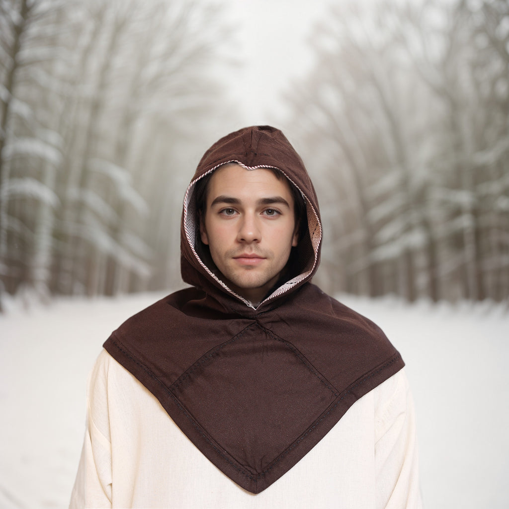 A young person wears a brown Reversible Viking Hood, Skjoldehamn, with a cream garment while standing on a snowy path lined with bare trees. The softly blurred background highlights the wintery medieval reenactment scene.