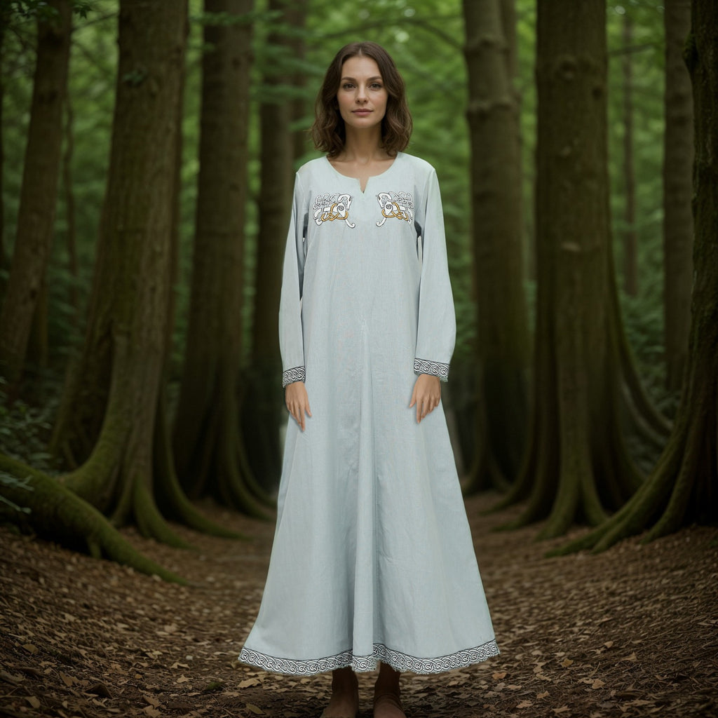 A woman stands barefoot on a forest floor, wearing the Sleipnir Viking Dress- Ladies—a long, light gray cotton-linen gown with embroidered details—surrounded by tall trees and lush green foliage in a tranquil woodland scene.