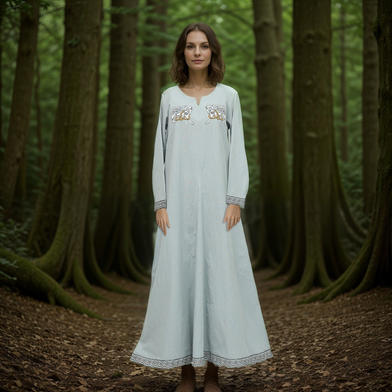 A woman wearing the Sleipnir Viking Dress- Ladies, a long light blue cotton-linen gown with embroidered patterns, stands barefoot on a mossy forest floor amid tall trees and lush foliage.