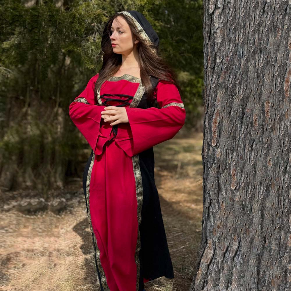 A woman wearing the Dark Majesty Medieval Dress stands by a tree in a wooded area, her hands folded and gaze thoughtful. Sunlight filters through the trees, highlighting her elegant LARP attire.