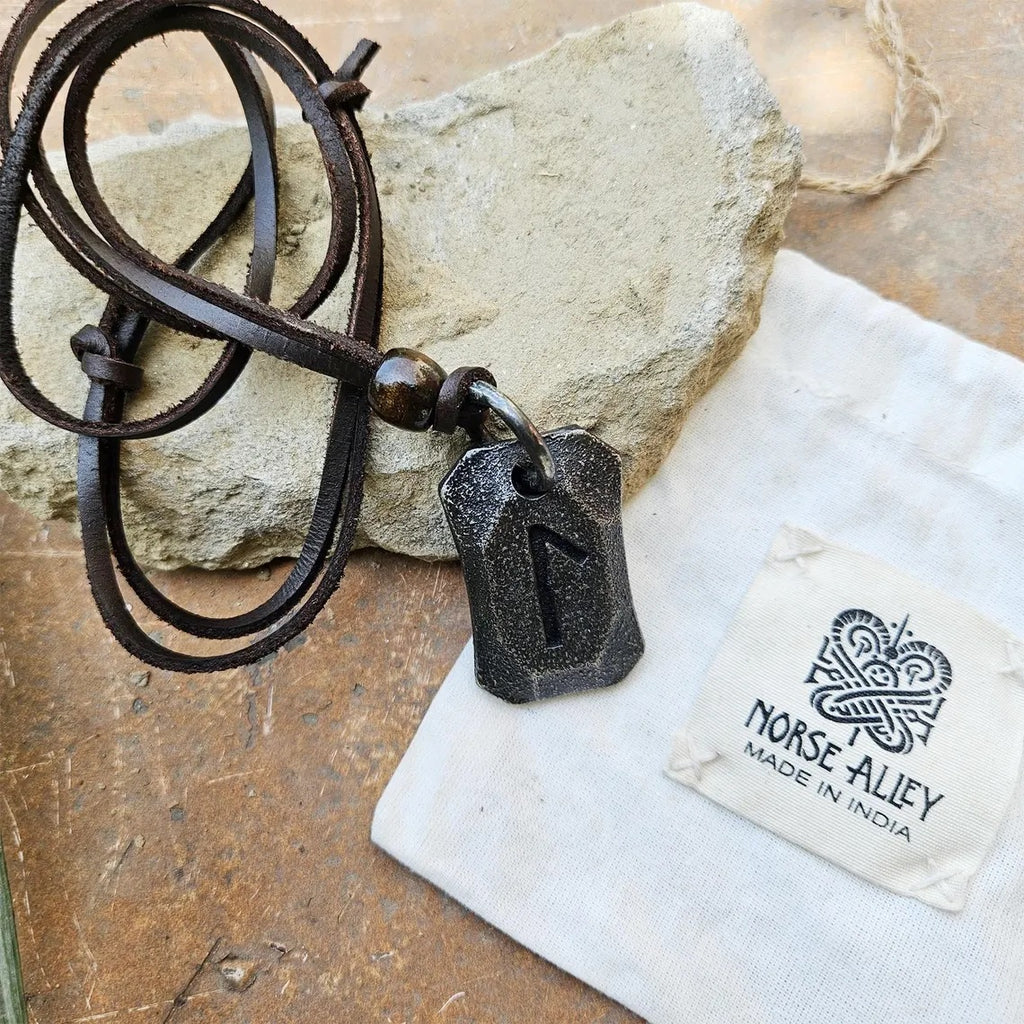 A black Viking Rune Pendant Laguz, inspired by Norse mythology, hangs from a leather cord on a rock. Next to it is a white pouch with the Norse Alley logo and "MADE IN INDIA" text.